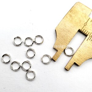 May include: A collection of small, shiny silver jump rings scattered on a white surface. A brass caliper is partially visible, positioned near some of the rings. The jump rings are circular and appear to be made of metal, likely for jewelry making or crafts.