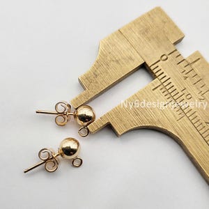 May include: Two gold-toned ball stud earrings with a push-back closure. The earrings are measured with a caliper, showing the size of the ball is approximately 6mm.