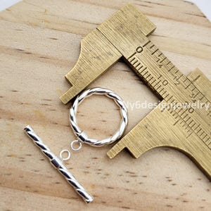 May include: A silver-colored toggle clasp with a twisted design, featuring a circular ring and a straight bar. The clasp is next to a brass measuring tool on a wooden surface. The text "Ny6designjewelry" is visible.