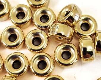 May include: A close-up of a group of gold-colored metal beads with a hole in the center. The beads are round and have a raised edge around the hole. The beads are arranged in a random pattern.