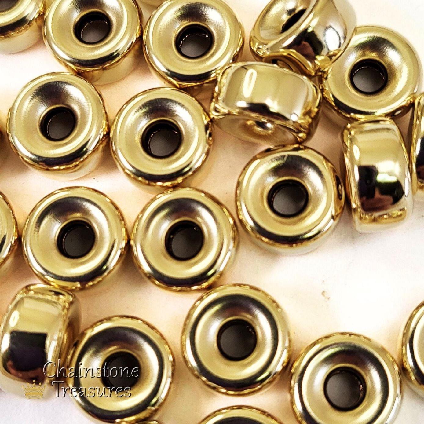 May include: A group of gold-colored metal beads with a hole in the center. The beads are round and have a raised edge around the hole. The beads are scattered on a white surface.