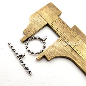 May include: Silver-toned metal toggle clasp set, featuring a twisted rope design. The set includes a circular clasp and a bar, both with snake head details. A brass caliper is in the background.