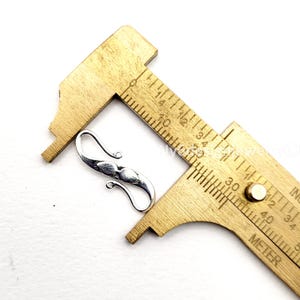 May include: A silver-colored hook clasp with a decorative design, next to a wooden ruler. The clasp is curved with a central, elongated shape. The ruler shows inch measurements.