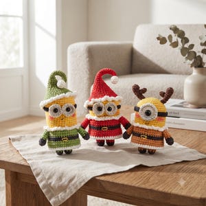 Peut inclure: Trois figurines crochetées ressemblant à des personnages cylindriques jaunes. La figurine de gauche porte un chapeau et une tenue verts avec une bordure blanche. Celle du centre porte un bonnet de Père Noël rouge. Celle de droite a des bois bruns.