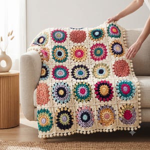Peut inclure: Une couverture en crochet faite à la main composée d'une grille de motifs carrés, chacun présentant un motif floral circulaire coloré dans des tons de rose, jaune, bleu et sarcelle, assemblés par du fil crème avec un pompon décoratif sur les bords.