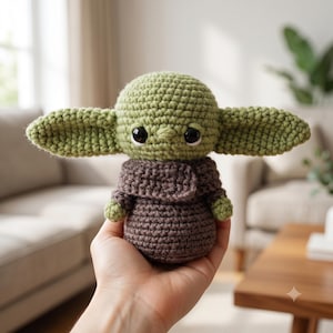 Peut inclure: Une petite poupée crochetée à la main avec une tête ronde verte, de grands yeux noirs et de longues oreilles pointues. La figurine porte une robe texturée marron foncé avec un col replié et de petites mains vertes visibles sur les côtés.