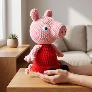 Peut inclure: Une peluche crochetée à la main représentant un personnage de cochon avec une grosse tête ronde, une texture en laine rose, deux oreilles, un museau et un œil blanc avec une pupille noire. La figurine porte une robe rouge vif et une queue frisée.