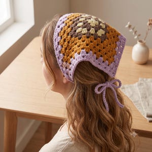 Peut inclure: Un foulard triangulaire en crochet avec un motif de granny square dans des tons crème, marron et jaune moutarde, bordé d'une bordure lavande clair. Le foulard comprend deux fines attaches en crochet lavande nouées à l'arrière.