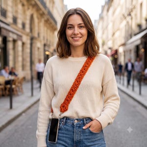 Peut inclure: Une longue sangle en macramé orange brûlé, épaisse et texturée, fixée à un smartphone dans une coque transparente. La sangle est portée en bandoulière sur un pull à manches longues couleur crème et un jean bleu en denim.