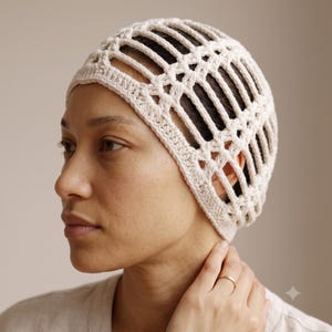 Peut inclure: Un bonnet en crochet de couleur crème présentant un motif en treillis ajouré avec des ouvertures rectangulaires verticales. Le couvre-chef est fabriqué en fil avec une bordure solide le long du bas, offrant une coupe structurée.