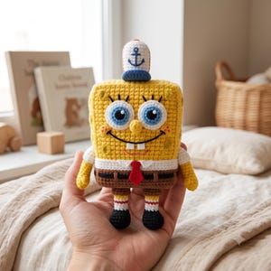 May include: A yellow rectangular crochet plush toy resembling a cartoon character with large blue eyes, a wide smile, and two teeth. It wears a white shirt with a red tie, brown pants, and a small white and blue cap with an anchor symbol on top.