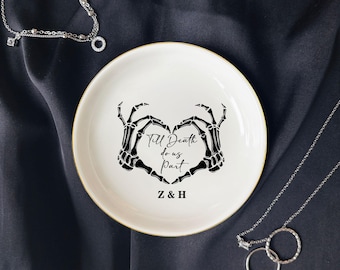 May include: A white ceramic trinket dish with a gold rim. The dish features a black skeleton hand heart design with the text "Till Death do us Part" and initials "Z & H". The dish is on a dark background with jewelry.