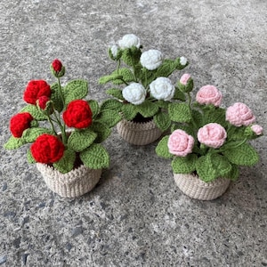 May include: Three crocheted potted plants with flowers. The plants are in brown pots and have green leaves. The flowers are red, white, and pink.