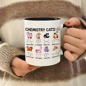 Könnte beinhalten: Ein weißer Keramikbecher mit schwarzem Henkel und schwarzem Innenraum. Die Außenseite zeigt den Text CHEMISTRY CATS und ein Atomsymbol, gefolgt von acht bunten Katzenillustrationen mit chemischen Strukturen und Beschriftungen: CHAOS CHEM, LIL SCIENTIST, NERDY CAT, BRAINY KITTY, SMART FLOOF, TINY GENIUS, LAB CHAOS und SCIENCE BABY.