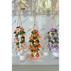May include: Three crocheted hanging flower pots with different colored flowers. The pots are made of beige yarn and the flowers are pink, orange, and purple.