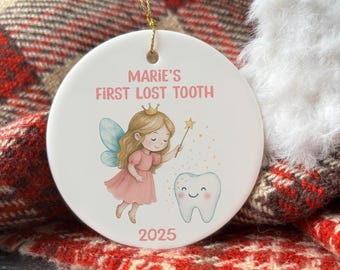 Può includere: Ornamento rotondo in ceramica bianca con cordino dorato per appenderlo. Presenta un'illustrazione di una fata con un vestito rosa e ali blu, un dente e la scritta "Marie's First Lost Tooth 2025". Lo sfondo è un motivo a quadri rossi e bianchi.