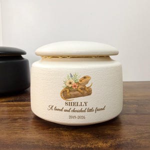 May include: A cream-colored ceramic urn with a lid, featuring a printed illustration of a bearded dragon on a log surrounded by flowers. The text reads SHELLY, A loved and cherished little friend, and the years 2019-2026 in dark brown lettering.