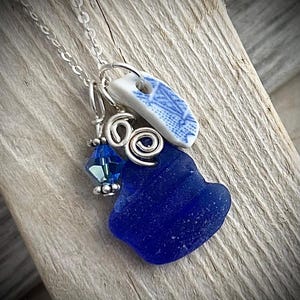 May include: A handcrafted necklace featuring a vibrant blue sea glass pendant, a small piece of blue and white ceramic, and a blue crystal bead. The pendant is suspended from a delicate silver chain, creating a unique and artistic accessory.
