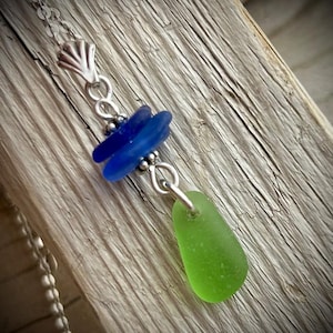 May include: A silver necklace featuring sea glass. The pendant has two blue sea glass pieces and one green sea glass piece. The necklace chain is silver and the clasp is a seashell design. The background is a wooden surface.
