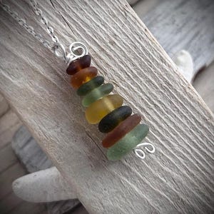May include: A necklace featuring a vertical stack of smooth, frosted sea glass pieces in shades of brown, olive green, and yellow. The glass segments are held together by a silver-toned wire wrap that forms small decorative coils at both ends.