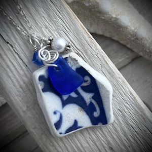 May include: A necklace featuring a pendant made from a piece of white and blue patterned ceramic pottery shard, accented with a small cobalt blue sea glass fragment, a white pearl bead, and a silver-toned metal wire spiral on a silver chain.