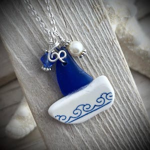 May include: A silver chain necklace featuring a pendant shaped like a sailboat, constructed from a triangular piece of cobalt blue sea glass and a white ceramic base with blue wave patterns, accented by a small blue crystal and a white pearl bead.