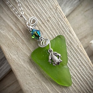 Peut inclure: Collier artisanal avec un pendentif en verre de mer vert vif. Le pendentif est orné d'un petit charm en forme de grenouille argentée et d'un fil décoratif avec un cristal vert et bleu. Le collier est suspendu à une chaîne argentée.