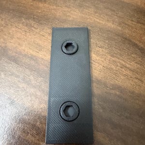 Puede incluir: Un soporte rectangular negro con dos pernos de cabeza hexagonal empotrados. El soporte tiene una superficie texturizada y probablemente está hecho de plástico. Los pernos están centrados y espaciados uniformemente.