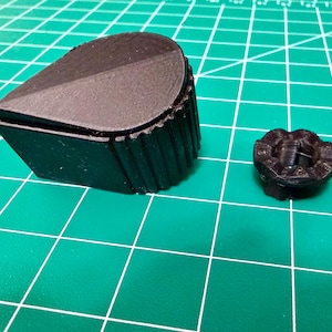 May include: Two black 3D-printed objects on a green grid surface. One is a large, angled, rectangular shape with a rounded top. The other is a small, circular object with a detailed, textured design.