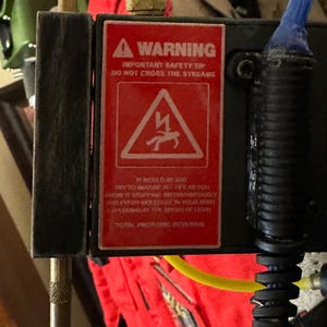 Può includere: Un cartello di avvertimento rosso con un fulmine e una figura stilizzata che cade. Il cartello recita "WARNING IMPORTANT SAFETY TIP DO NOT CROSS THE STREAMS IT WOULD BE BAD TRY TO IMAGINE ALT THE AS YOU KNOW IT STOPPING ISTANTANEOUTRY AND I'VERY MORE CULY IN YOUR CORRY EXPLODING AT THE SPEED OF LIGHE TOTAL PROTONIC REVERSAL"