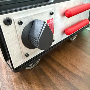 May include: A close-up of a silver and black machine with red cylindrical handles. A large black knob and a warning label are visible on the front panel. The machine has small wheels and is set on a wooden surface.