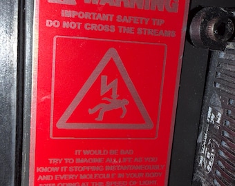 Puede incluir: Una etiqueta de advertencia rectangular roja con borde negro que presenta un icono de rayo blanco y una figura humana. El texto dice: WARNING IMPORTANT SAFETY TIP DO NOT CROSS THE STREAMS IT WOULD BE BAD TOTAL PROTONIC REVERSAL.