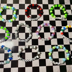 May include: A collection of eleven circular beaded bracelets made from colorful plastic pony beads. Each bracelet features white letter beads spelling out words including WEB, MALWARE, ONLINE, UPLOAD, INTERNET, PIXEL, WINDOWS 2000, CYBER, LOADING, and DIGITAL.