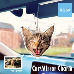 May include: A clear plastic car mirror charm featuring a cat's head with a playful expression. The charm is hanging from a string and has the text "Car Mirror Charm" printed below it. The image also includes a smaller image of a cat with the text "your photo" below it.