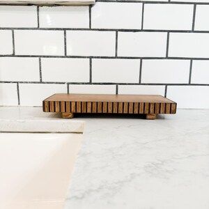 May include: A rectangular wooden serving tray with a slatted top and small feet. The tray is made of light brown wood and sits on a white countertop. The background features white subway tile.
