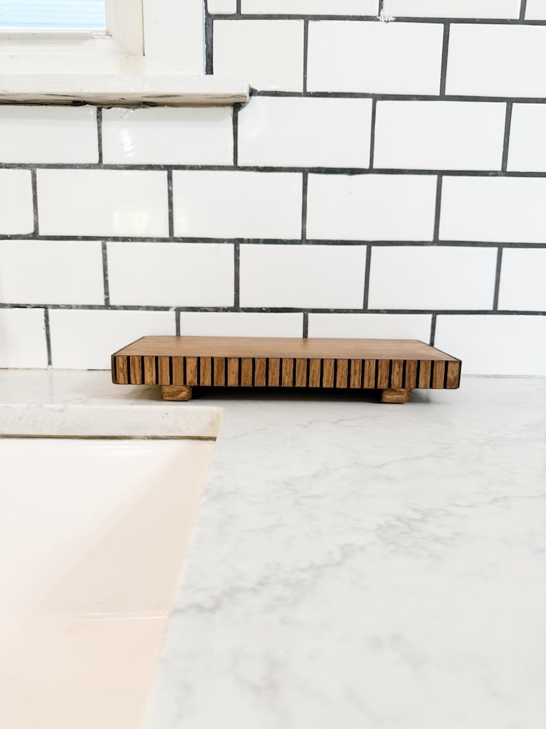 May include: A rectangular wooden serving tray with a slatted top and small feet. The tray is made of light brown wood and sits on a white countertop. The background features white subway tile.