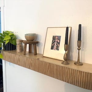 May include: A light wood floating shelf with a vertical fluted front edge. On the shelf sits a green plant in a black pot, a small wooden stool with a ceramic bowl, a gold-toned picture frame containing a photograph, and two brass candle holders.