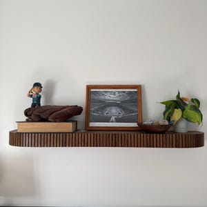 May include: A dark brown, ribbed wooden shelf displays a baseball bobblehead, a brown baseball glove, a framed black and white stadium photo, a small wooden bowl, and a potted plant. The shelf is mounted on a white wall above a blue paneled wall.