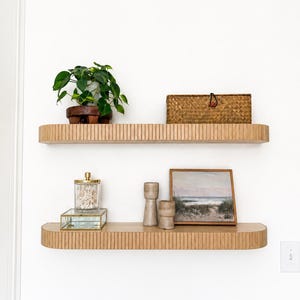 May include: Two light wood floating shelves with a vertical ribbed front edge. The top shelf holds a green potted plant and a rectangular woven basket. The bottom shelf holds a glass jar, a small glass box, two ceramic vases, and a framed landscape painting.