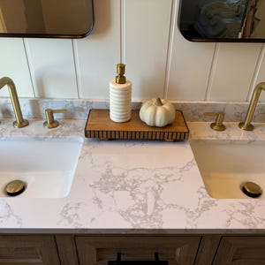 May include: A bathroom vanity with two white sinks and gold fixtures. A wooden tray holds a white and gold striped soap dispenser and a small white pumpkin. The countertop has a gray and white marble pattern.