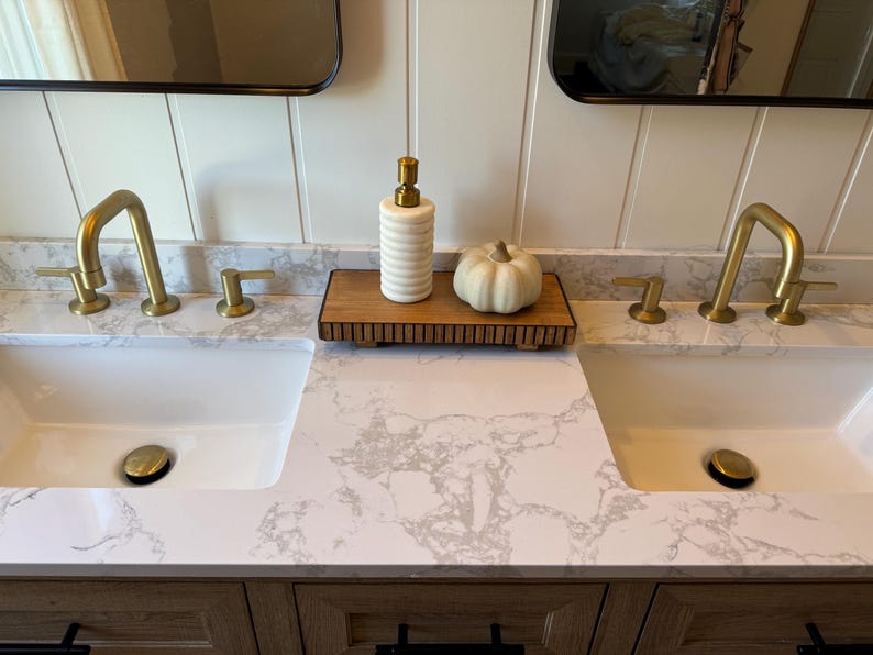 May include: A bathroom vanity with two white sinks and gold fixtures. A wooden tray holds a white and gold striped soap dispenser and a small white pumpkin. The countertop has a gray and white marble pattern.