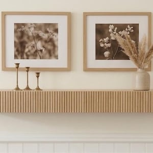 May include: A light wood floating shelf with a vertical slatted front design. Above the shelf are two framed sepia-toned botanical prints, three thin brass candlesticks, and a ceramic vase containing dried pampas grass stems.