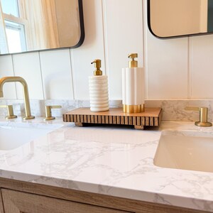 May include: Bathroom vanity with a marble countertop, two white sinks, and gold fixtures. Two white and gold soap dispensers sit on a wooden tray. The vanity has a light wood finish and black drawer pulls. Two black framed mirrors are visible.