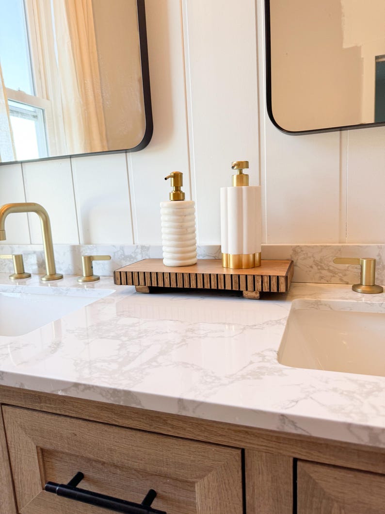 May include: Bathroom vanity with a marble countertop, two white sinks, and gold fixtures. Two white and gold soap dispensers sit on a wooden tray. The vanity has a light wood finish and black drawer pulls. Two black framed mirrors are visible.