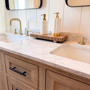 May include: Bathroom vanity with two sinks, gold faucets, and black hardware. Two white soap dispensers with gold pumps sit on a wooden tray. The vanity has light wood cabinets and a marble countertop. Mirrors with black frames are visible.
