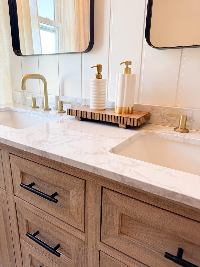 May include: Bathroom vanity with two sinks, gold faucets, and black hardware. Two white soap dispensers with gold pumps sit on a wooden tray. The vanity has light wood cabinets and a marble countertop. Mirrors with black frames are visible.