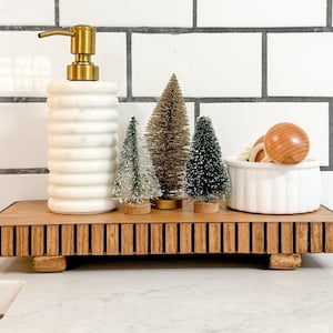 May include: A decorative wooden tray with a ribbed design holds a white soap dispenser with a gold pump, miniature Christmas trees, and a small white bowl with wooden accents. The background features white subway tile.