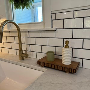 May include: A wooden tray with a ribbed design holds a green ceramic cup and a white and gold soap dispenser. The tray sits on a countertop next to a white sink and a gold faucet. White subway tile is in the background.