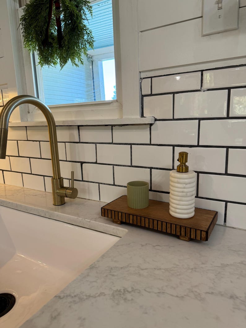 May include: A wooden tray with a ribbed design holds a green ceramic cup and a white and gold soap dispenser. The tray sits on a countertop next to a white sink and a gold faucet. White subway tile is in the background.