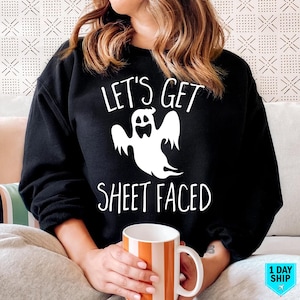 May include: Black sweatshirt featuring the phrase "LET'S GET SHEET FACED" and a white ghost design. The person is holding a white and orange striped mug. This is a Halloween-themed garment.